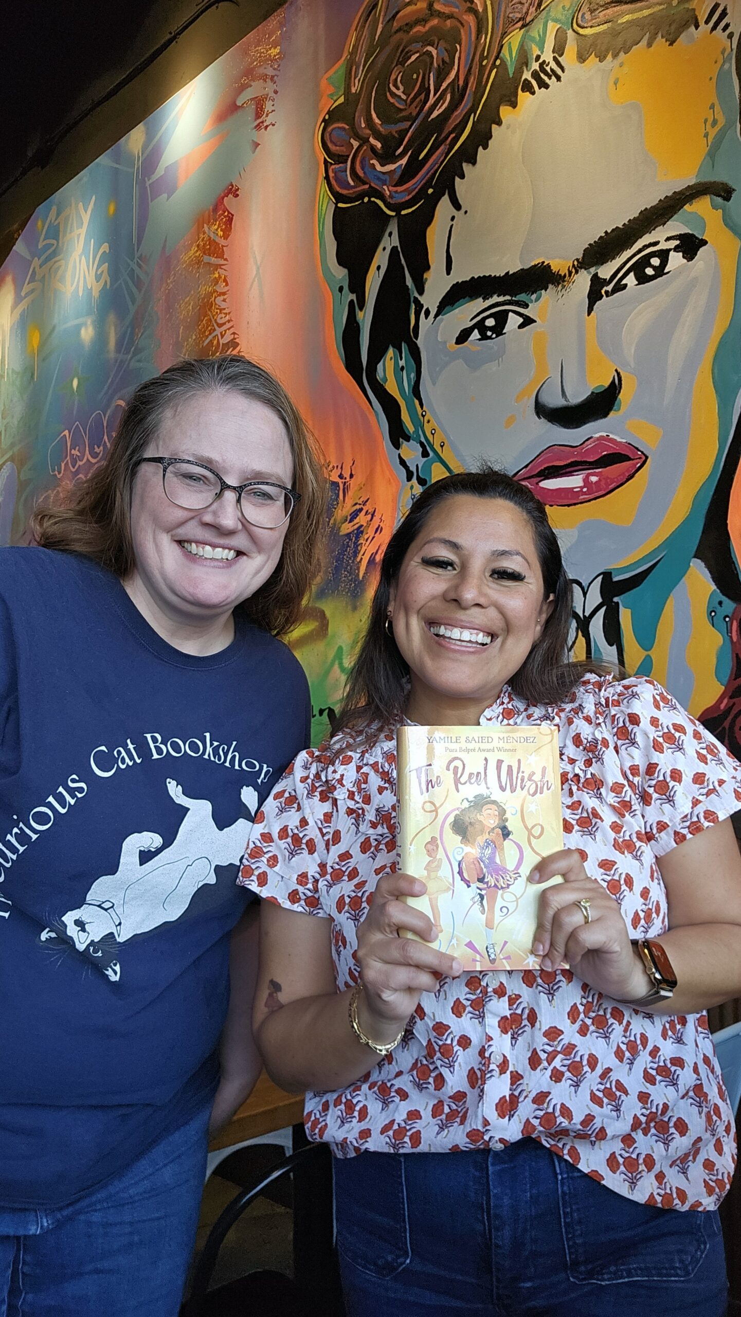 Stacy, a pale woman with long light hair and glasses, stands next to Yamile, a brown-haired, brown-skinned woman who is smiling and holding a copy of The Reel Wish, which Yamile wrote and Stacy edited. They stand in front of a mural of the face of Frida Kahlo.